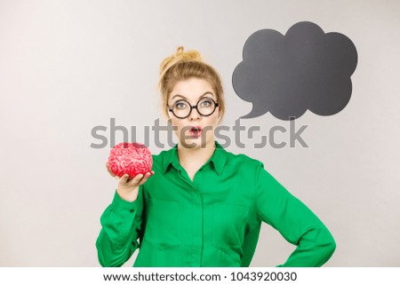 Business woman wearing green jacket and eyeglasses intensive thinking finding great problem solution holding fake brain, black thinking or speech bubble next to her.