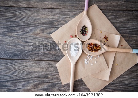 Horizontal table top shot of 3 wooden spoons facing each other, filled with course salt, pepper and dried chili flakes. Spoons are resting on brown paper and shot against a wooden textured background