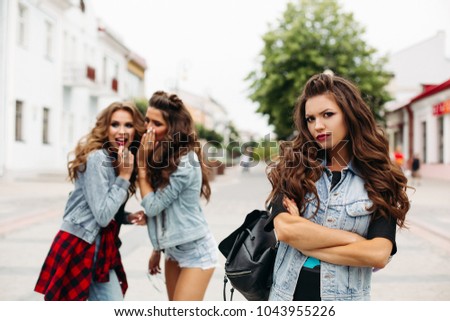 Two trendy teen girls with hairstyles wearing denim clothes gossiping about third stading in foreground with folded arms and looking at camera with worried look. City street.