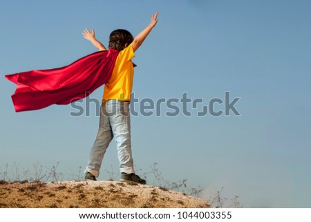 Boy playing superheroes on the sky background, teenage superhero in a red cloak on a hill