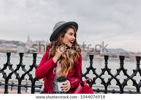 Attractive woman with wavy hair standing at bridge and looking at river in cloudy day. Outdoor portrait of fashionable young lady in hat posing with cup of coffee and enjoying city views.