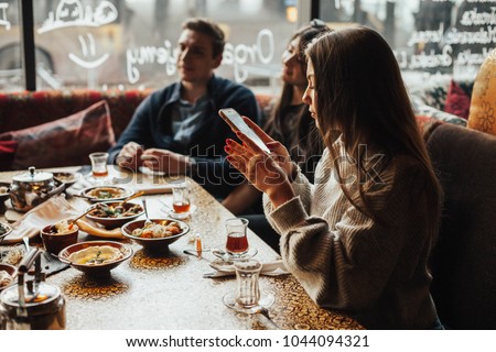 Young girl is taking pictures of food. A young company of people is smoking a hookah and communicating in an oriental restaurant. Lebanon cuisine served in restaurant.  Traditional meze lunch