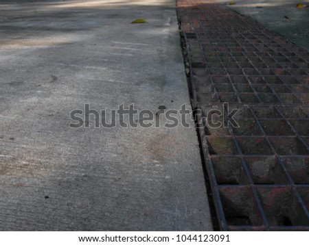 Sewage drain separating road and walkway. Concrete ground and pavement. Worn out rusted metal grate.