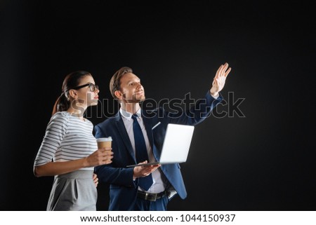 Show you something. Pleasant handsome unshaken man standing in the dark room with his colleague holding the laptop holding hand up.