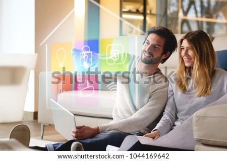 A couple sitting on the sofa controls all the functions of the house such as wi-fi, heating, lighting, television through holography. Concept of, home automation, automations, future, technology. Royalty-Free Stock Photo #1044169642