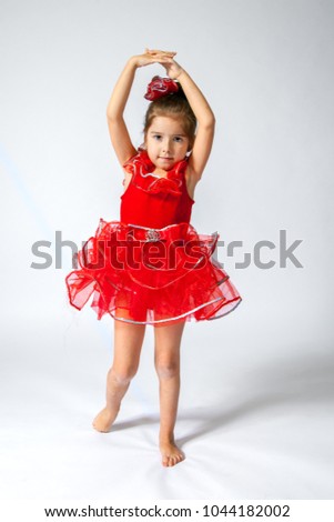 A sweet, little girl in a red ballet costume poses with hands above her head on a white background.   She is attempting fifth position.  She is isolated with a path.