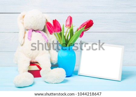Colorful red spring tulip flowers in nice blue vase, blank photo frame and stuffed toy bunny on light wooden background as greeting card. Mothersday or spring concept