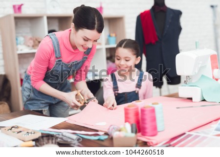 A woman cuts out a piece of clothing from the fabric. Next to her is a little girl. They are with Mom in the sewing workshop.