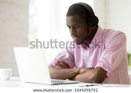 African american man wearing headphones using laptop looking at computer screen, smiling young black businessman learning studying online, watching video or making call with pc internet application Royalty-Free Stock Photo #1044298792