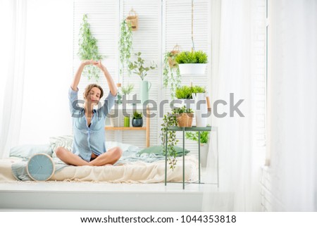 Young woman having a good morning sitting on the bed in the beautiful bright bedroom with green plants and clock Royalty-Free Stock Photo #1044353818