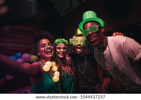 Group of friends enjoying at party in the bar and taking selfie with smart phone. Cheerful en and women partying and celebrating St.Patrick's Day at night club.