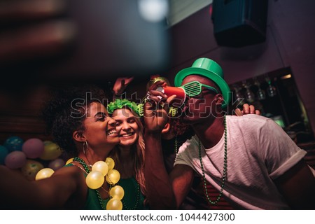 Man drinking a glass of beer with female friends smiling and taking selfie at bar. Young people celebrating St.Patricks day at night club and taking selfie with phone.