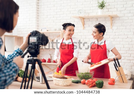Two culinary bloggers in red aprons mixing diced peppers with salad to camera.
