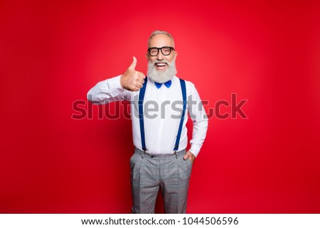 Portrait of cheerful, laughing old barber, stylist showing thumb up holding hand in pocket having blue suspenders, pants, bowtie, isolated on red background