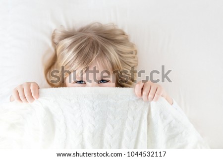 Little girl peeking out blanket on bed. Cute kid smiling and hiding under knitted cover. Palyful and mischievous eyes. Hide-and-seek. Children having fun playing active games. Happy childhood concept