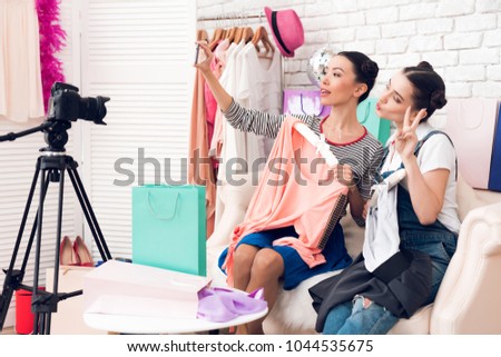 Two fashion blogger girls in jeans and shirt with skirt hold up two colorful dresses to camera taking selfie.
