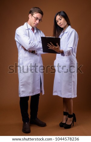 Studio shot of young handsome man doctor and young beautiful Asian woman doctor together against brown background
