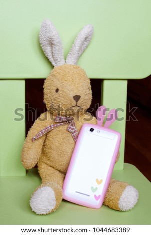 Light brown bunny toy sitting on green chair and holding white phone with pink bunny ears as birthday gift. Happy Easter, mothers, fathers day or birthday greeting card with space for text on phone.