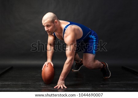 A young athletic man in blue wrestling tights and blue shorts stands in a low position and holds a brown rugby ball on a black isolated background in a photo studio