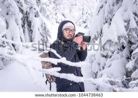 A young man European appearance in glasses with a backpack and a camera in the hands of a winter forest picks up