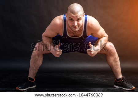 A young athletic man in blue wrestling tights and blue shorts makes a squat with a wide leg arrangement and shows thumbs up on a black isolated background in a photo studio