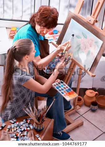 Artist painting easel in studio. Authentic grandmother and kids girl paints with palette watercolor paints and brush morning sunlight. Students learn older woman to draw. Individual family training.