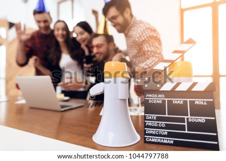 Young office workers looking at laptop screen together. They have glasses with champagne in their hands. They are in a good mood. In the foreground is a speakerphone and a movie clapboard