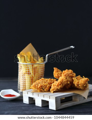 Crispy chicken strips with delish spicy ketchup in ramekin and french fries in a fancy support, wooden plate. Dark background. Junk fast food concept. Selective focus. Copy space for text Royalty-Free Stock Photo #1044844459