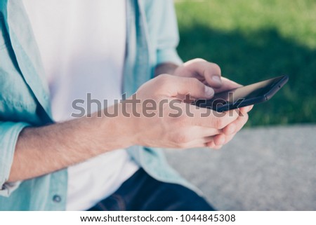 Modern technology people remote work job video concept. Cropped close up photo of handsome man's hands typing message on new stylish smartphone wearing jeans denim casual shirt green grass