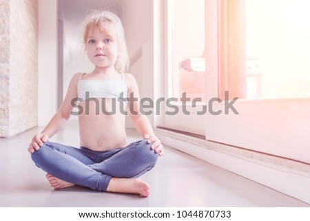 Little blond girl in white top and blue leggings doing a yoga exercise in the sunny room