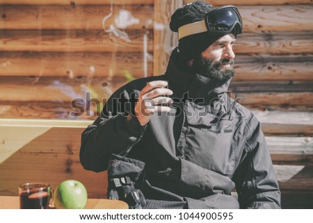 Bearded cool snowboarded taking rest after ride session.Young man drinking cup of hot tea on sunny terrace. Blurred background.Horizontal.Visual effects