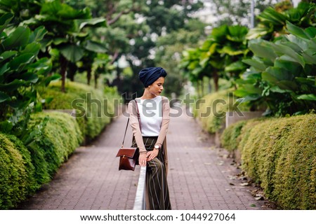 Portrait of a young Muslim woman in a hijab leaning on a railing in the park. She is wearing a blue turban and is elegantly dressed; she is of Arab, Malay, Asian descent and is attractive.  Royalty-Free Stock Photo #1044927064
