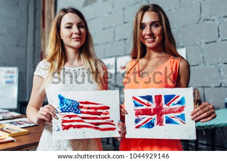 Close-up image of two young women holding a drawing of British and American flags hand-drawn with aquarelle technique on plain paper