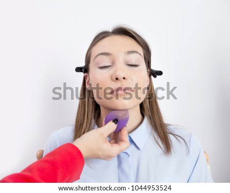 A make up artist is applying powder with a soft purple sponge under her client's chin. The client's eyes are closed.