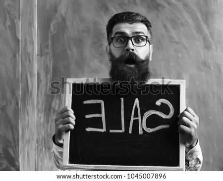 Bearded man, long beard. Brutal caucasian unshaven surprised hipster with glasses and moustache holding sale inscription on blackboard on brown studio background
