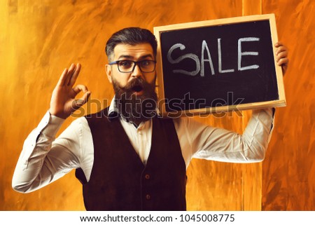 Bearded man, long beard. Brutal caucasian unshaven surprised hipster with glasses and moustache holding sale inscription on blackboard wearing white shirt, suede waistcoat on brown studio background