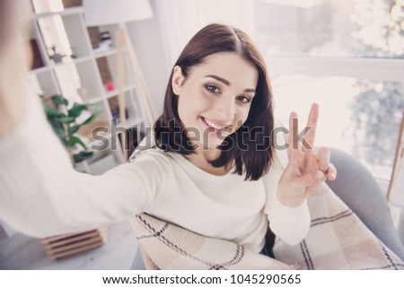 Self portrait of pretty, charming, nice, cute, stylish, brunette girl in pullover with modern hairstyle, shooting selfie gesturing v-sign to the front camera, wrapped in plaid, sitting in living room