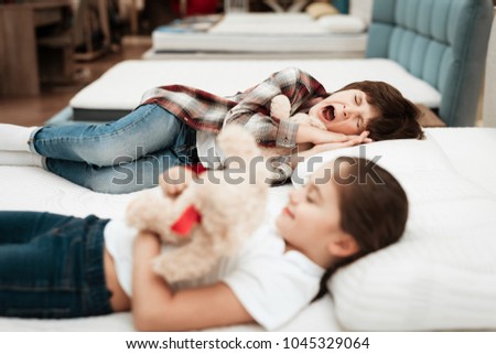 Little happy girl hugs bear lying on the bed. In the background a little boy sleeps. Cute girl yawns, hugging teddy bear on mattress in the store.