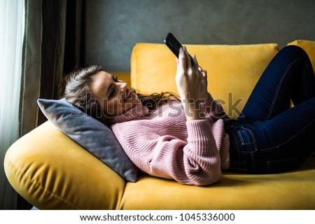 Close up of a relaxed girl using a smart phone lying on a sofa in the living room at home with a cozy background Royalty-Free Stock Photo #1045336000