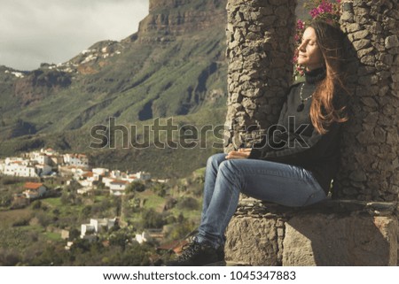 Relaxed young woman sitting in park with great views of valley village and green mountains on the background. Pretty lady resting while enjoying the sun outdoors. Tourist sunbathing in nature