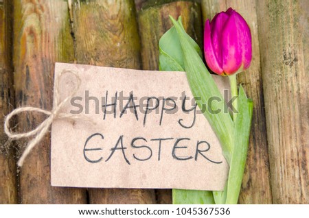 Purple tulip flower and carton paper with handwriting message: happy Easter. Wooden fence wall background.