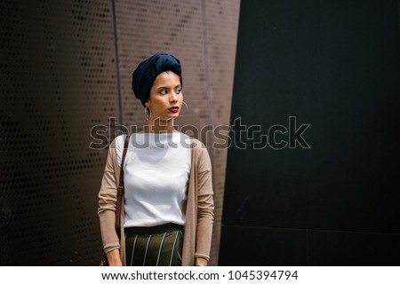 Portrait of a Muslim woman wearing a turban (hijab, head scarf) in a studio, against a metal background. She is elegantly dressed and Saudi Arabian, Malay, Asian descent.  Royalty-Free Stock Photo #1045394794