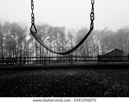 Black and white of a lone empty swing on an dreary cold foggy morning with a barn and a wooden fence in the background. Royalty-Free Stock Photo #1045396870