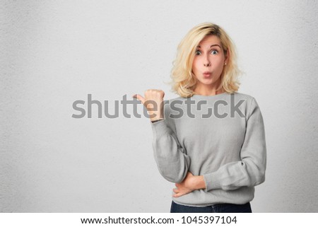 Portrait of attractive blonde girl with pierced nose indicates with thumb aside, wears gray sweater, being shocked. copy space for your promotional text