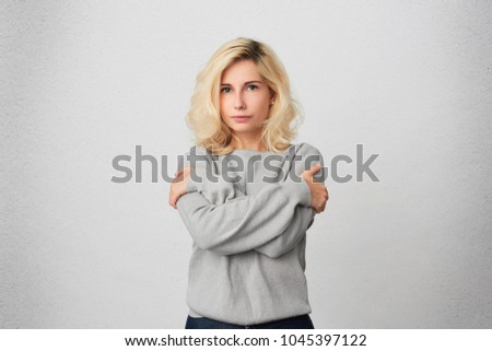 Portrait of young blonde girl embrace herself, wears casual gray sweater and feels comfortable and warm. Negative emotions. Isolated over white wall
