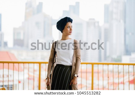 Portrait of a young, attractive and elegant Muslim woman wearing a turban (hijab, head scarf, tudung). The backdrop is a city skyline of Singapore's business district. She is of Malay or Arab descent. Royalty-Free Stock Photo #1045513282