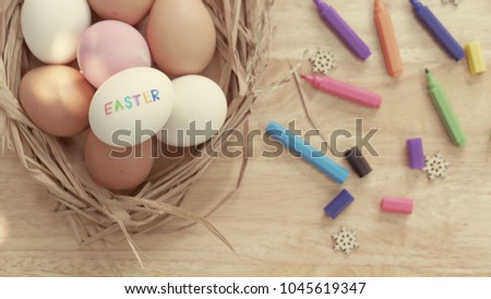 Many white, pink and orange eggs are in the straw basket and put on the wooden table with many colorful pens. The picture concepts are food, Easter Card, painting, healthy, organic in vintage style.