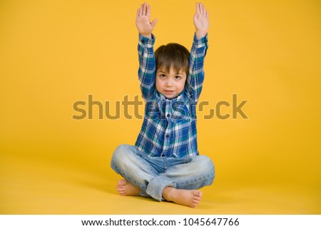 Portrait of boy in blue shirt and jeans on yellow background sitting cross-legged, hands up.