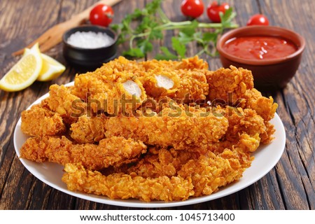 close-up of delicious crispy fried chicken breast strips on white plate, on old rustic  wooden table with tomato sauce and lemon slices, easy recipe for outdoor picnic or party, view from above Royalty-Free Stock Photo #1045713400