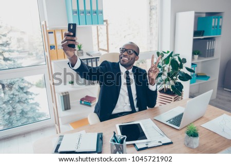 Stylish attractive, modern, cheerful man shooting self picture on front camera of his telephone, smart phone, showing peace symbol, two fingers, having video call, fun, sitting in work place, station